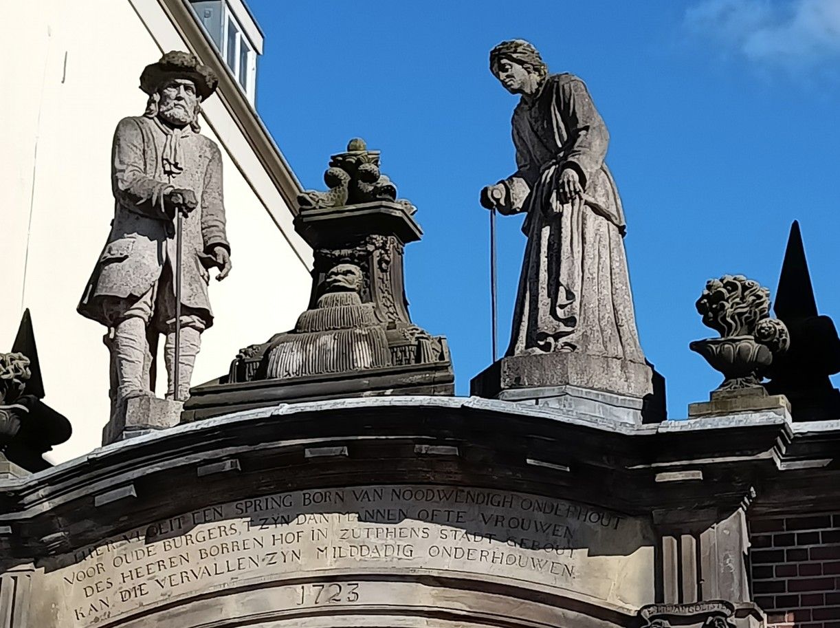 Beelden boven een boogvormige ingang, waaronder twee figuren en sierlijke houtsnijwerken, onder een blauwe hemel.