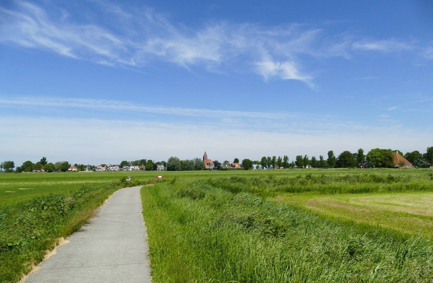 Een geplaveid pad door een groen veld leidt naar een dorp in de verte, onder een blauwe hemel.