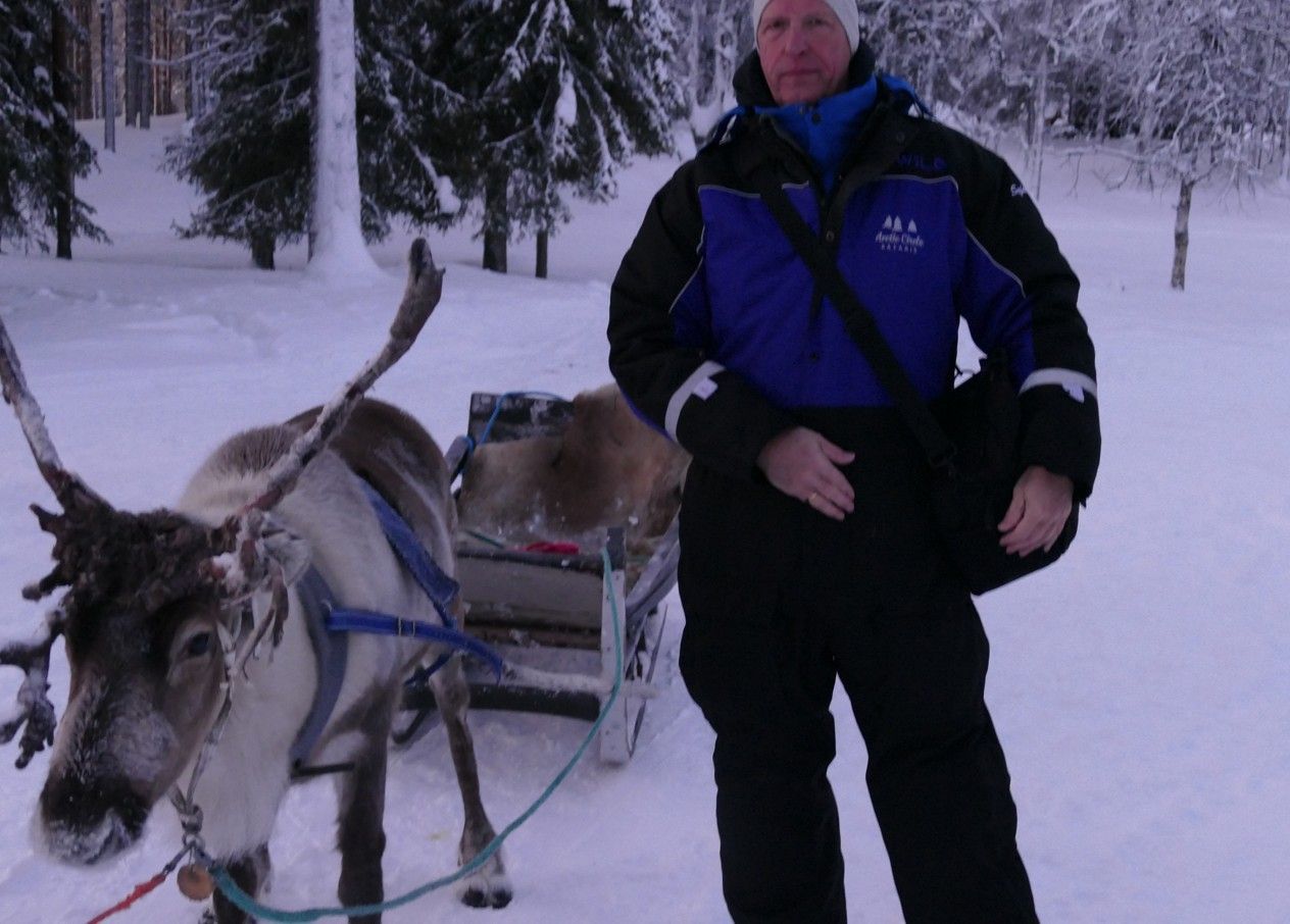Een man staat naast een rendier dat een slee trekt in een besneeuwd bos.
