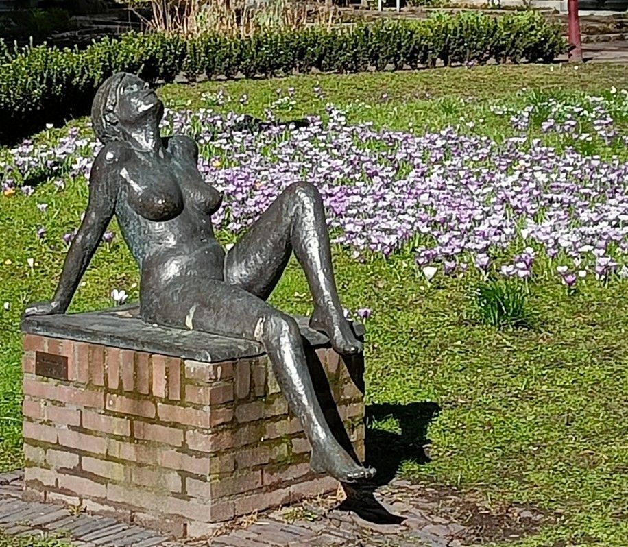 Bronzen beeld van een zittende vrouw, achteroverleunend met het hoofd achterover gekanteld, op een bakstenen sokkel, in een bloementuin.