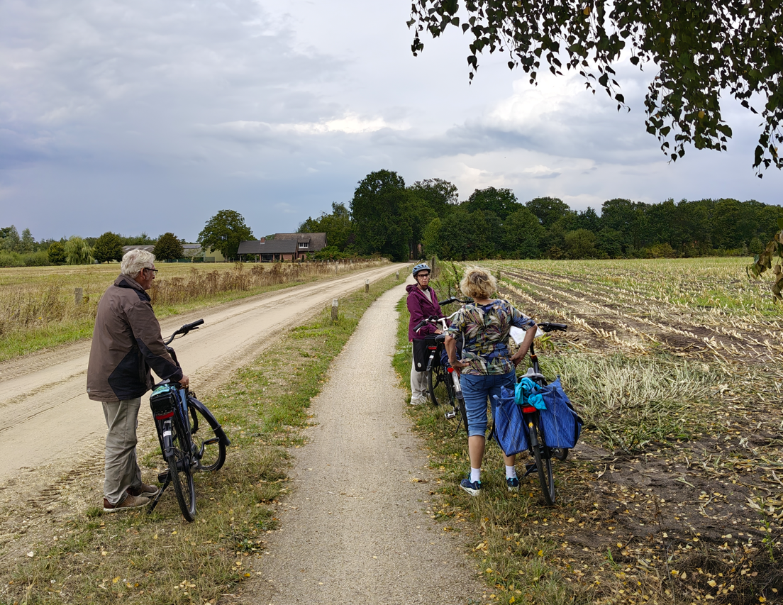 Fietsers met bepakte fietsen op een onverharde landweg langs geoogste velden onder een bewolkte hemel.