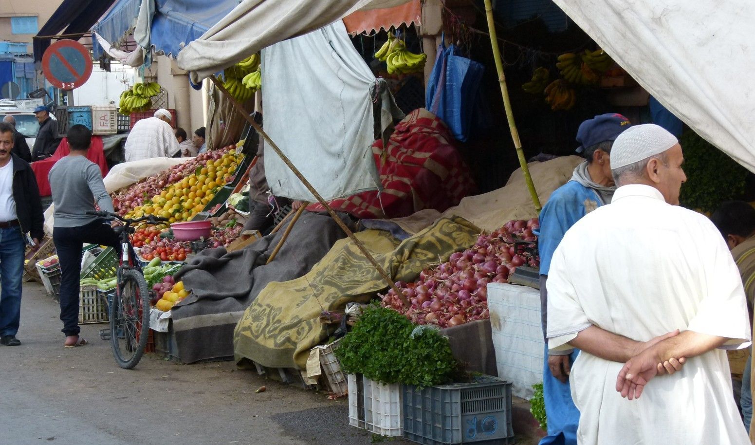 Straatmarkt met producten uitgestald onder luifels; mensen lopen er rond.