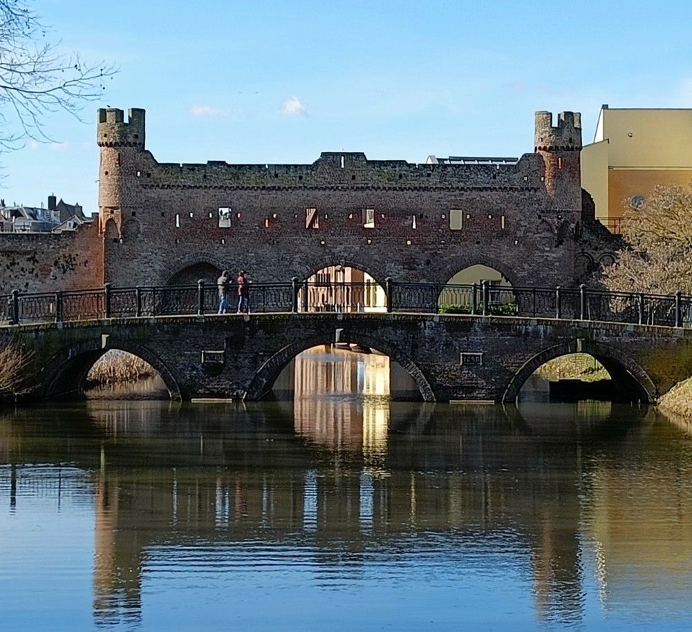 Een stenen brug met boogvormige openingen, die leidt naar een middeleeuws poortgebouw met torentjes boven een waterpartij.