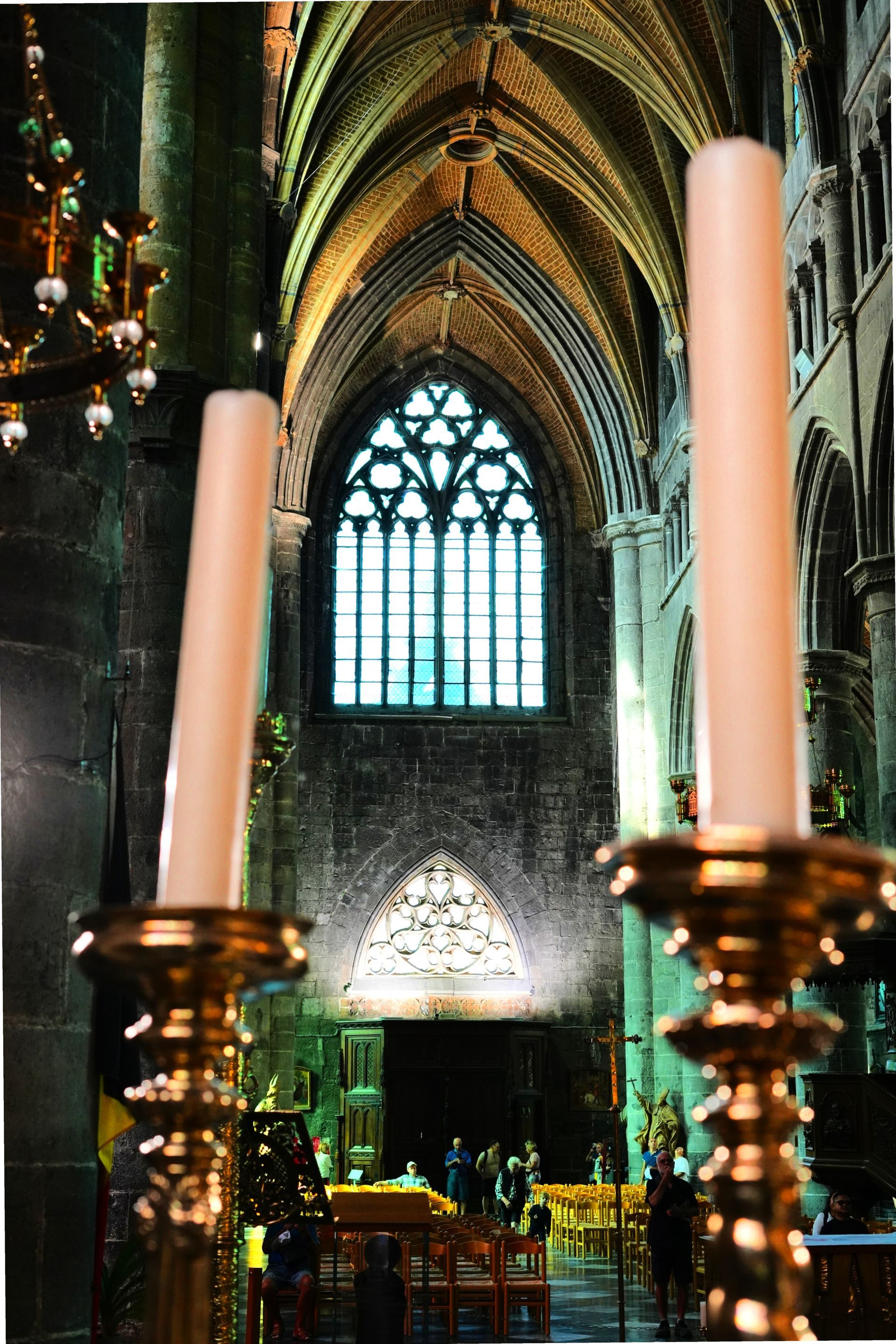 Kaarsen omlijsten het interieur van een gotische kathedraal met een glas-in-loodraam en een gewelfd plafond.