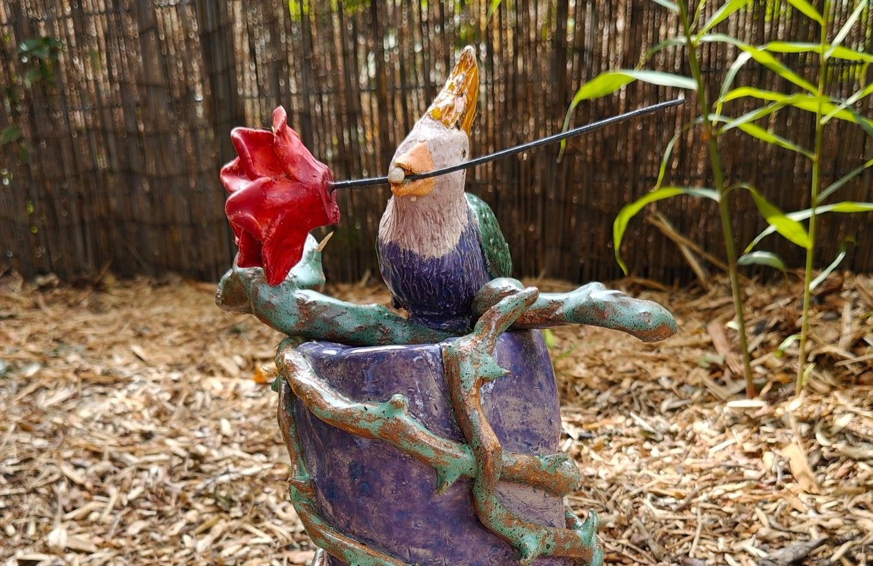 Kleurrijke vogelsculptuur met een rode bloem in een tuin met houtsnippers.
