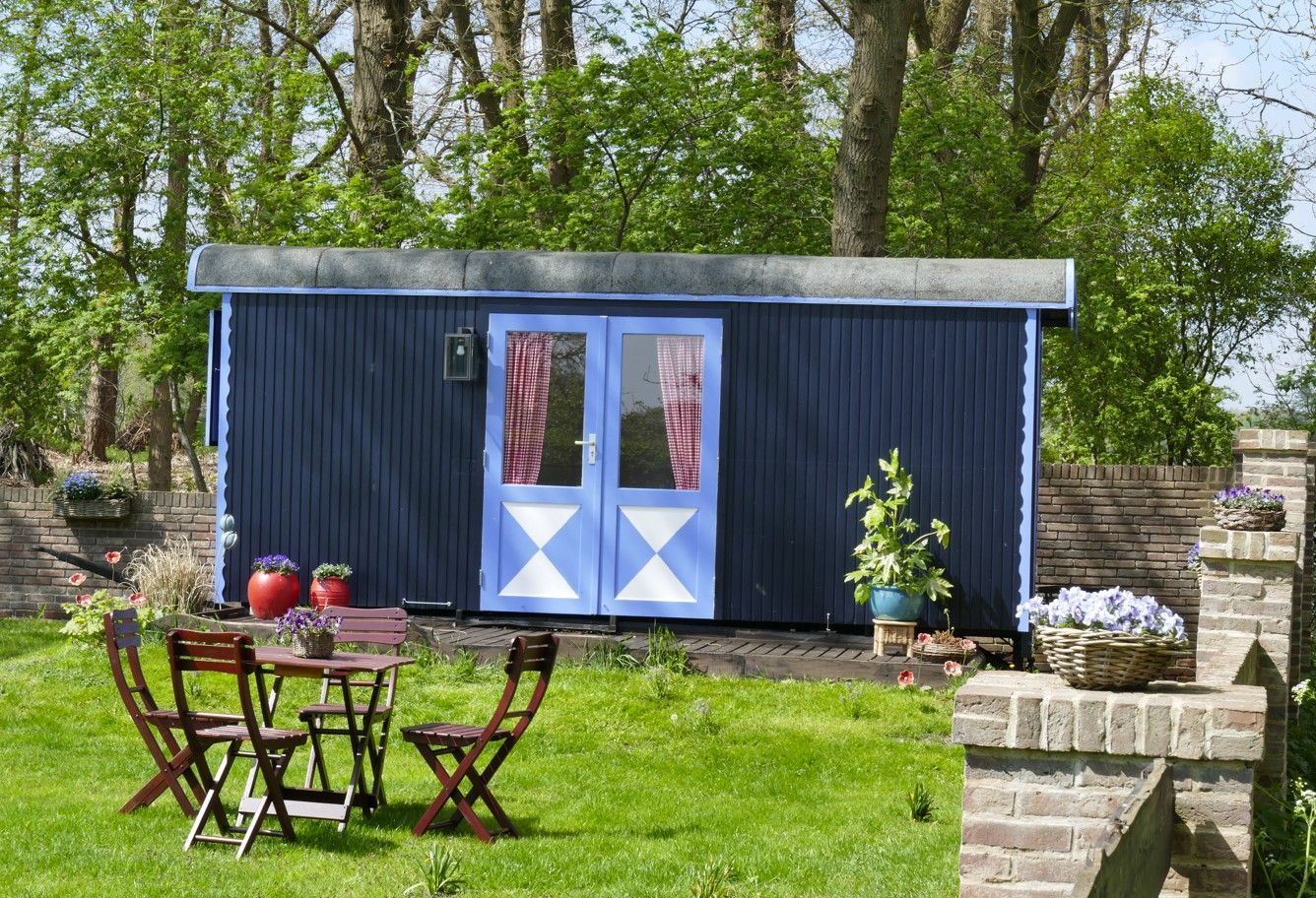 Blauwe schuur met dubbele deuren, rode gordijnen en witte ruitvormige accenten. Tafel en stoelen op het gazon.