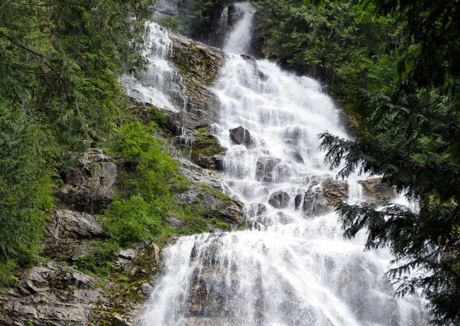 Een waterval die langs een rotsachtige berghelling naar beneden stort, omgeven door groene bomen.