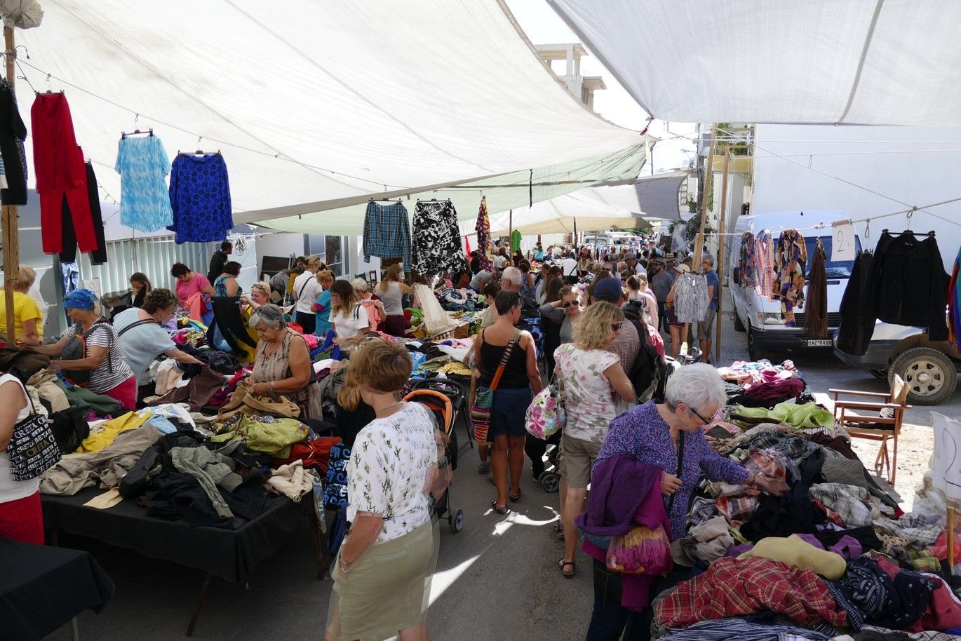 Een openluchtmarkt met veel winkelend publiek dat onder een witte luifel langs kledingrekken loopt.