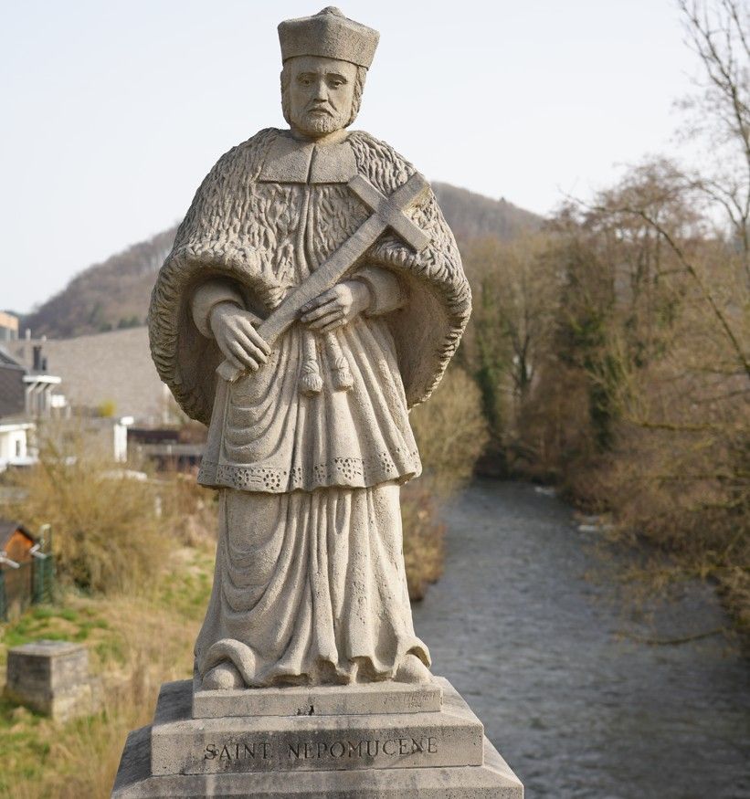 Een stenen beeld van een religieuze figuur die een kruis vasthoudt, staande op een brug over een rivier.
