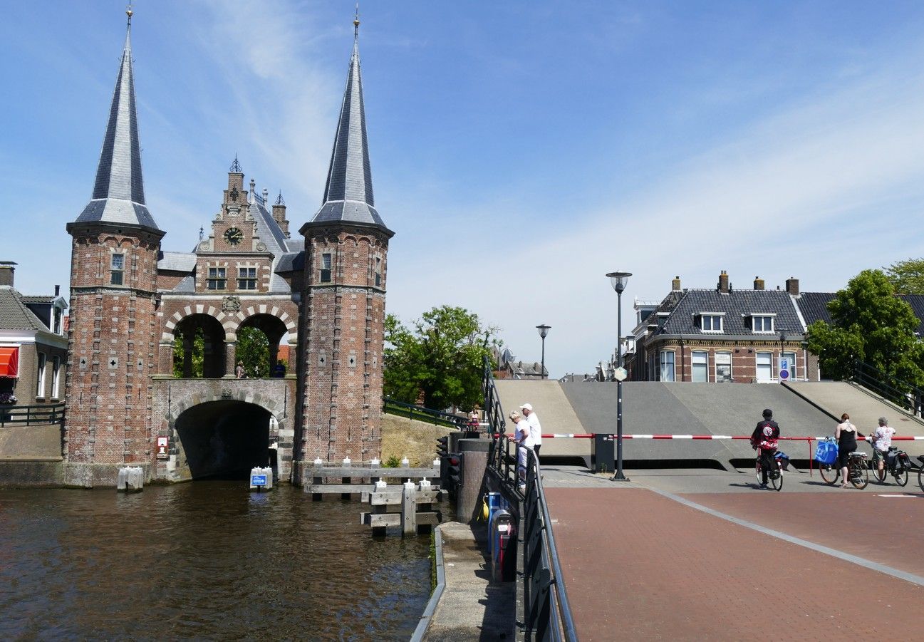 Historische waterpoort met twee torens, kanaal, rode bakstenen brug en blauwe lucht. Mensen bij de slagboom.
