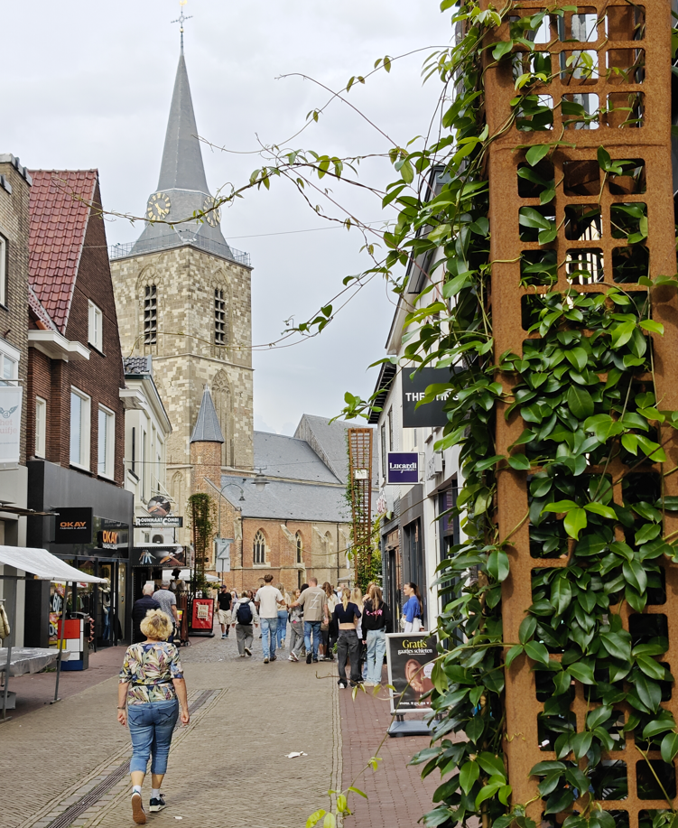 Een straat met kinderkopjes in een historische stad, met voetgangers en een kerktoren op de achtergrond.