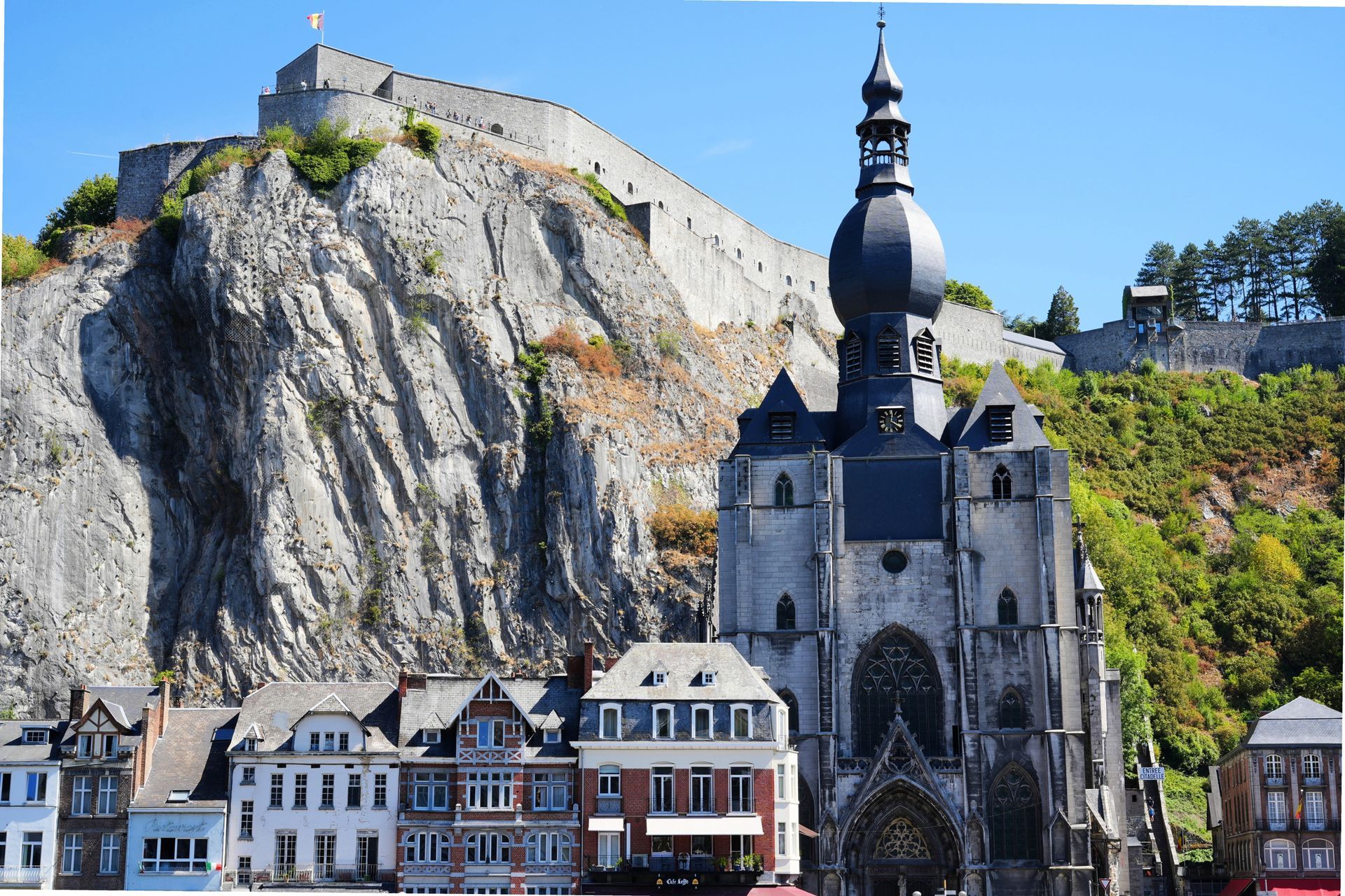 Dinant - Historische kerk onder een torenhoge klif, met kleurrijke gebouwen langs de rivier en het fort op de heuveltop.