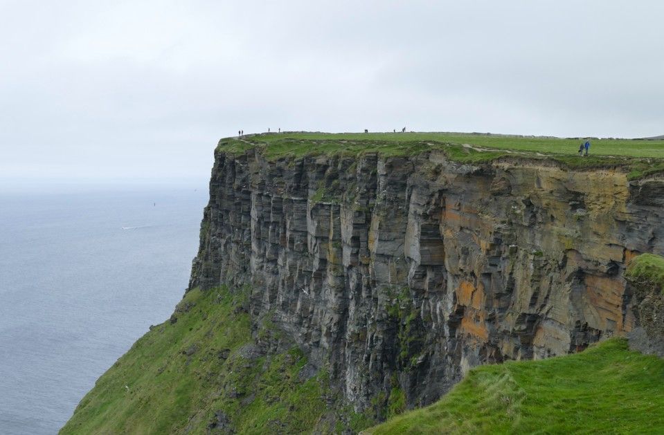 De kliffen van Moher in Ierland steken uit in de oceaan. Groen gras bovenop en aan de voet; een grijze rotswand. 
