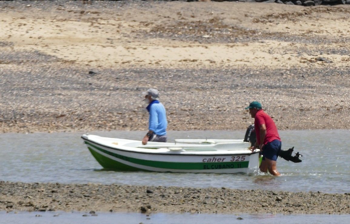 Twee mensen duwen een witgroene boot door ondiep water vlakbij een zandstrand.