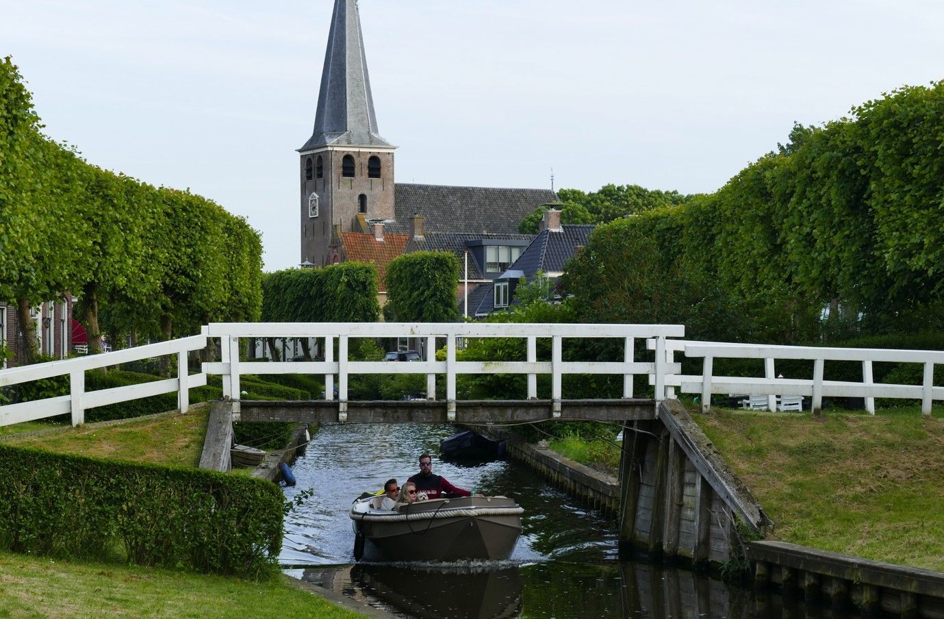 Een boot vaart onder een witte brug door, met een kerk op de achtergrond. Groene hagen en gras omzomen het water.