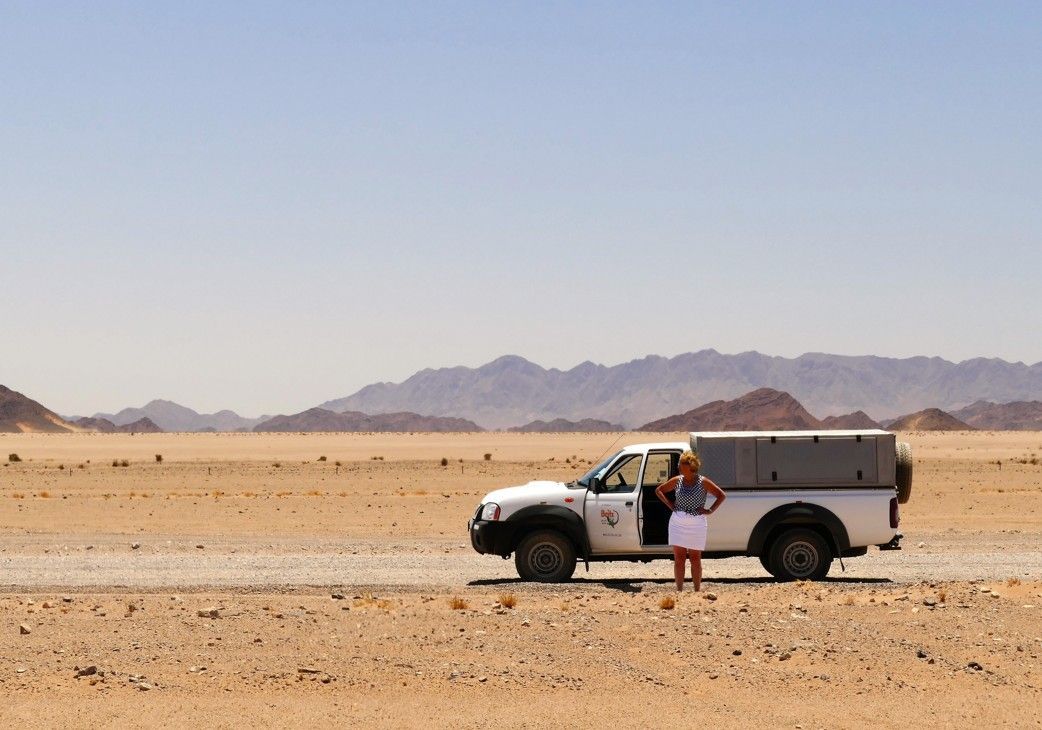 Vrouw staat naast een witte vrachtwagen op een woestijnweg, met bergen in de verte onder een blauwe hemel.