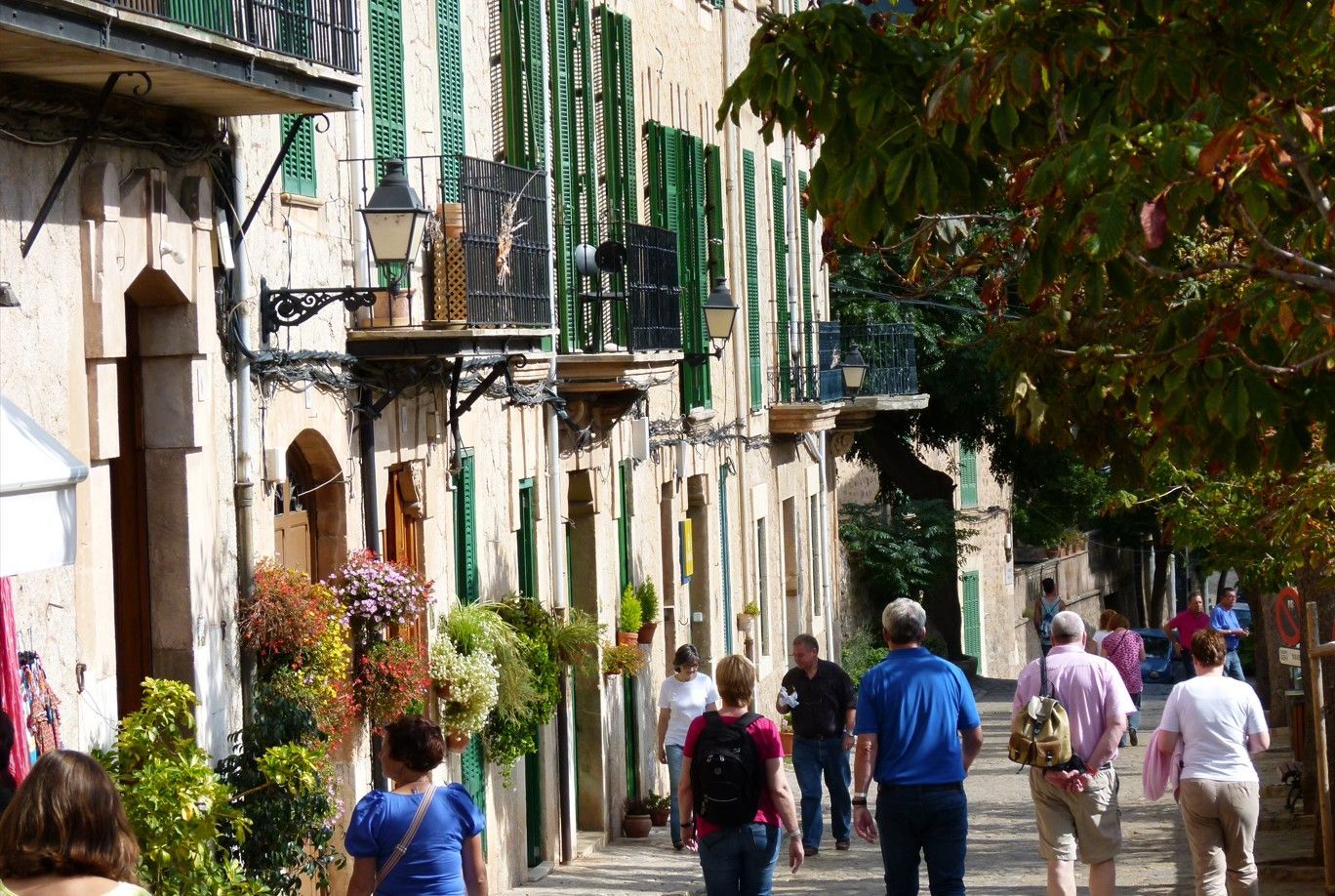 Smalle Europese straat met gebouwen; mensen wandelen in de zon. Groene luiken, bloembakken en balkons.