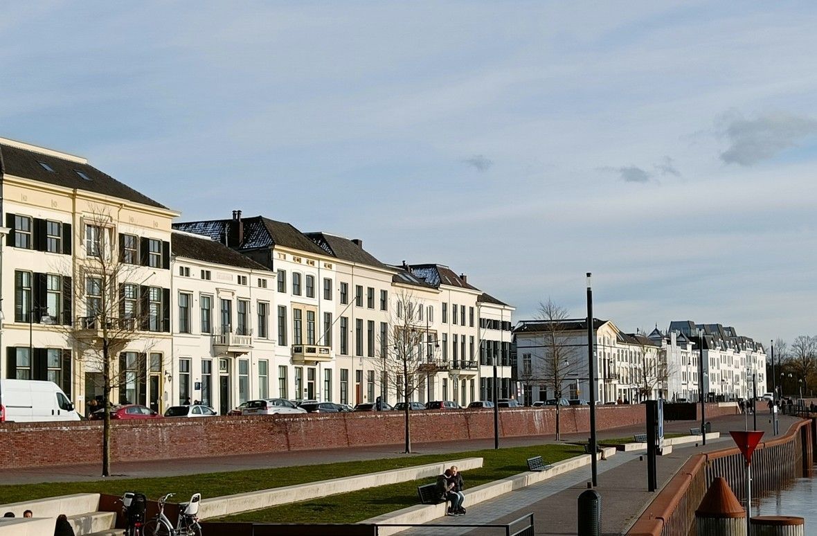 Een rij witte gebouwen met donkere luiken langs een promenade van rode bakstenen, onder een lichtblauwe hemel.