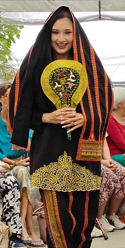 Vrouw in een traditionele zwart-oranje jurk met een decoratieve waaier in haar hand, staand in de Botanische tuin Delft