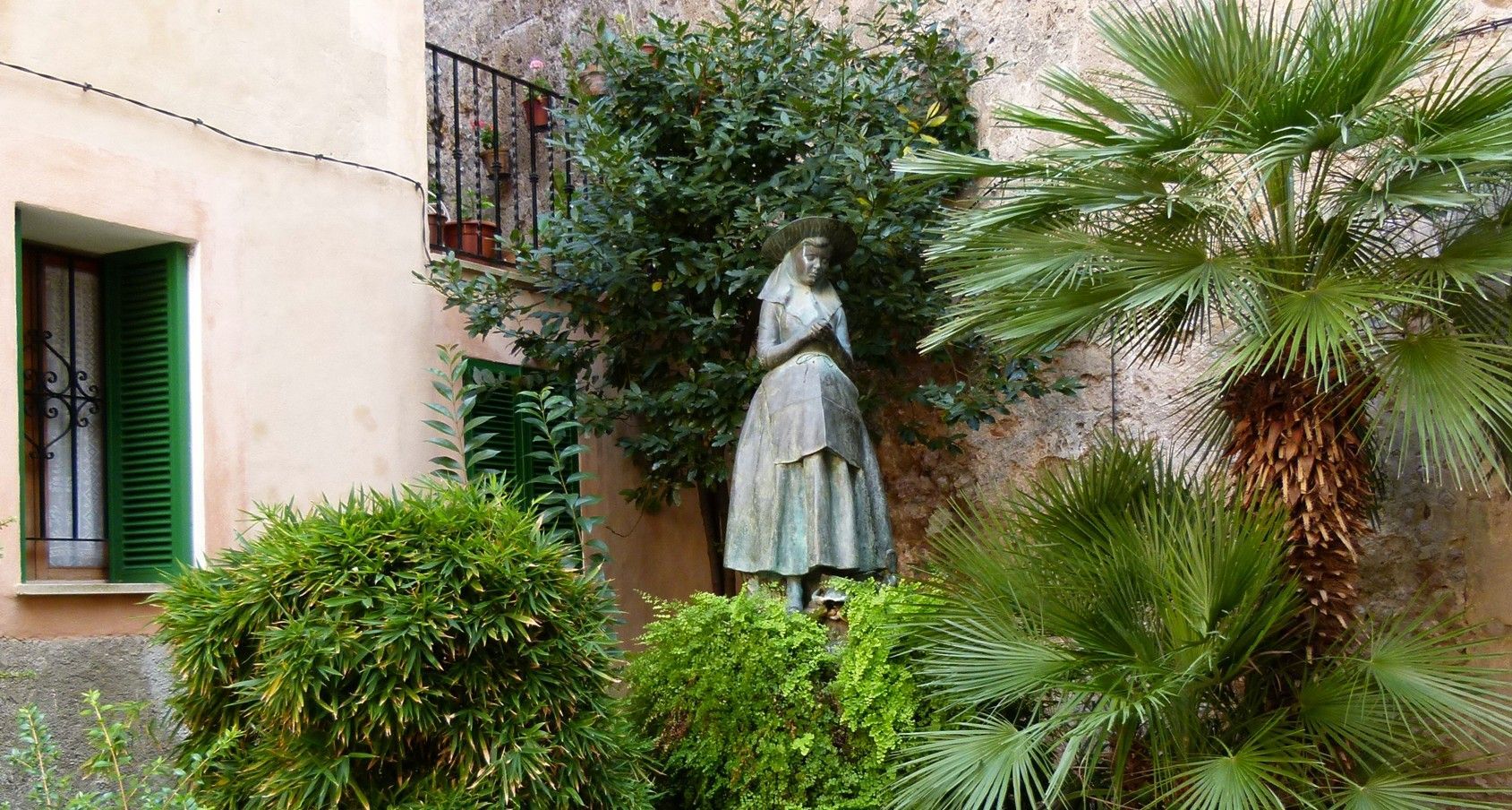 Standbeeld van een vrouw in een tuin met weelderig groen en een palmboom, beige gebouwen op de achtergrond.