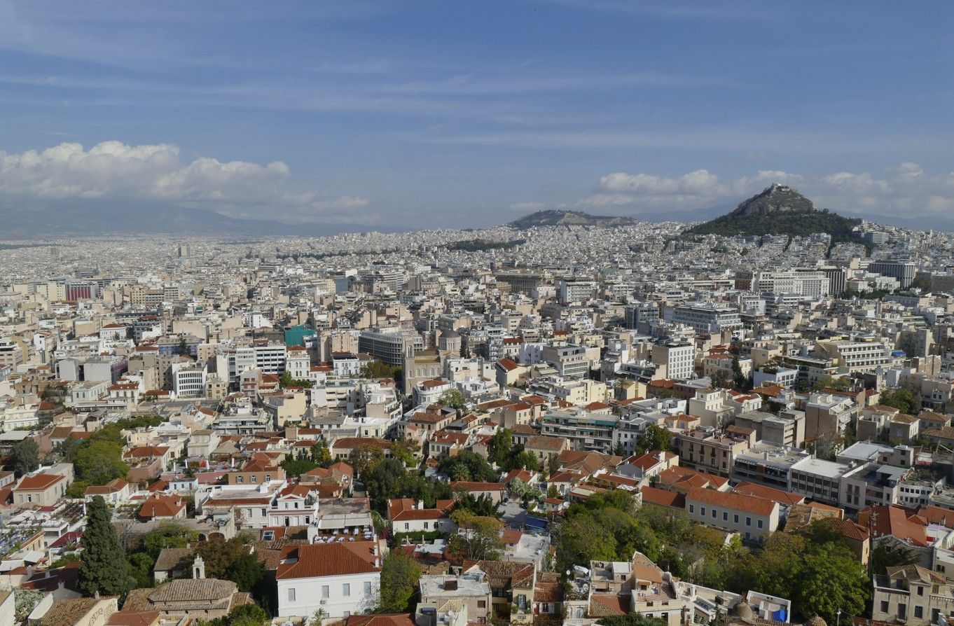 Panoramisch uitzicht op Athene, Griekenland. Gebouwen strekken zich uit over het landschap onder een blauwe hemel. In de verte is een heuvel te zien.