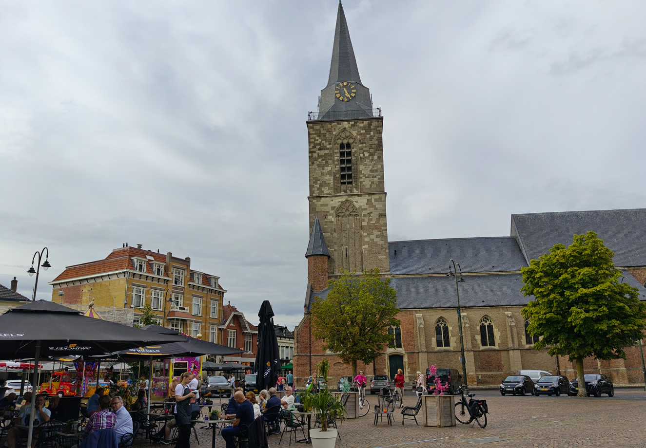 Stadsplein met een stenen kerktoren, marktkramen en mensen onder een bewolkte hemel.