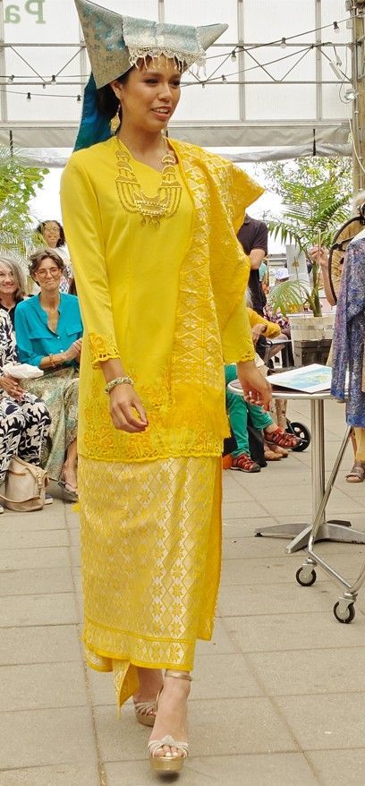 Een persoon in een felgele traditionele outfit loopt over een catwalk en draagt ​​een zilveren hoofdtooi.
