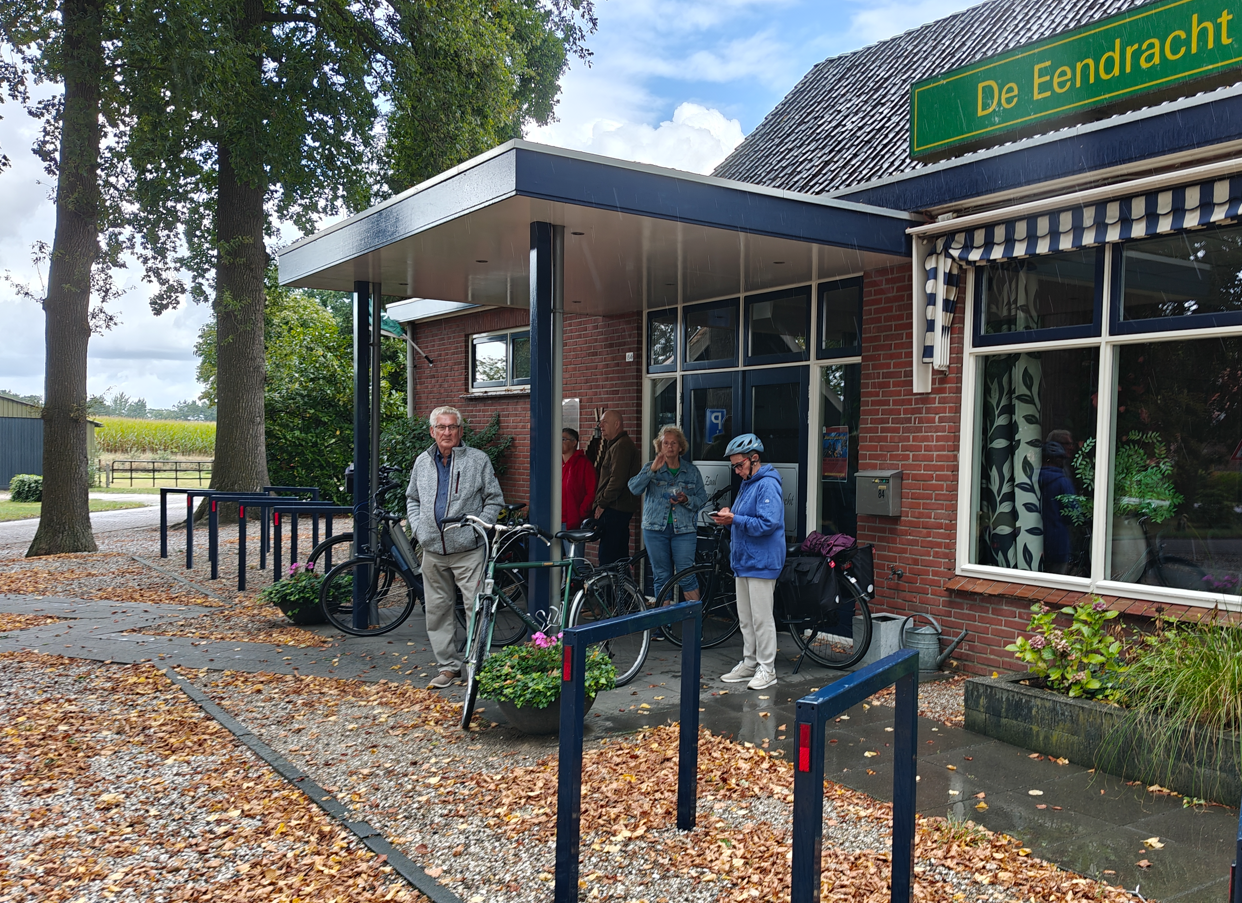Mensen met fietsen staan buiten een bakstenen gebouw met een groen bord, op een natte herfstmiddag.