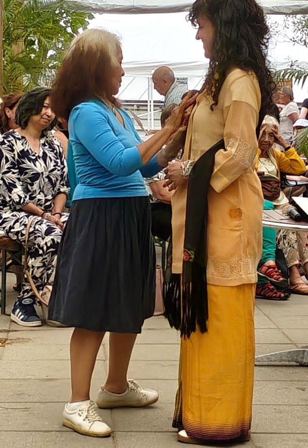 Twee vrouwen staan ​​buiten; de ene speldt iets vast op de beige jas en gele jurk van de andere.
