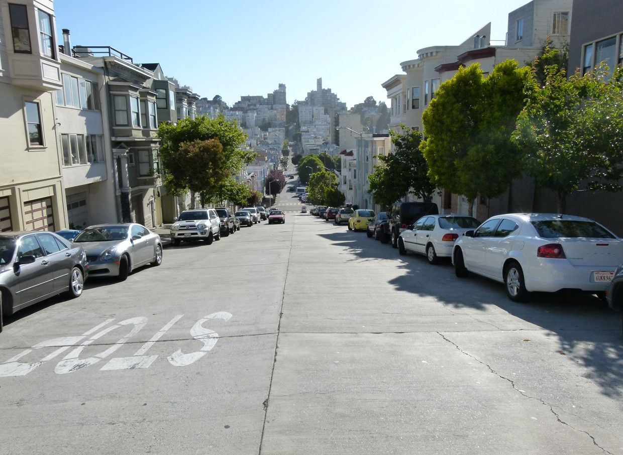 Een straat in San Francisco, vol geparkeerde auto's en gebouwen, loopt bergopwaarts richting de structuren in de verte.