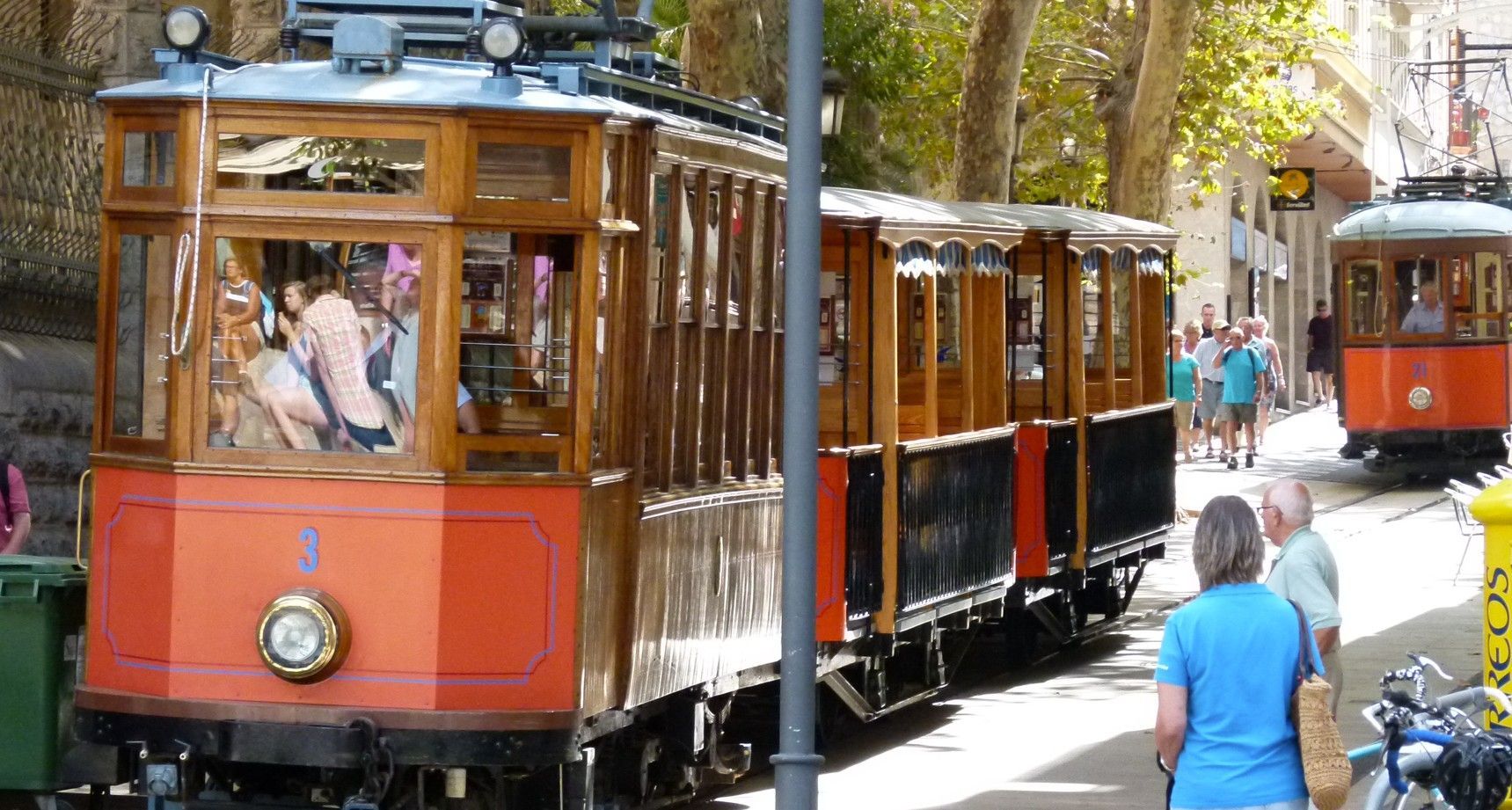 Houten trams, oranje en bruin geschilderd, rijden door de stad, met mensen die er vlakbij staan.