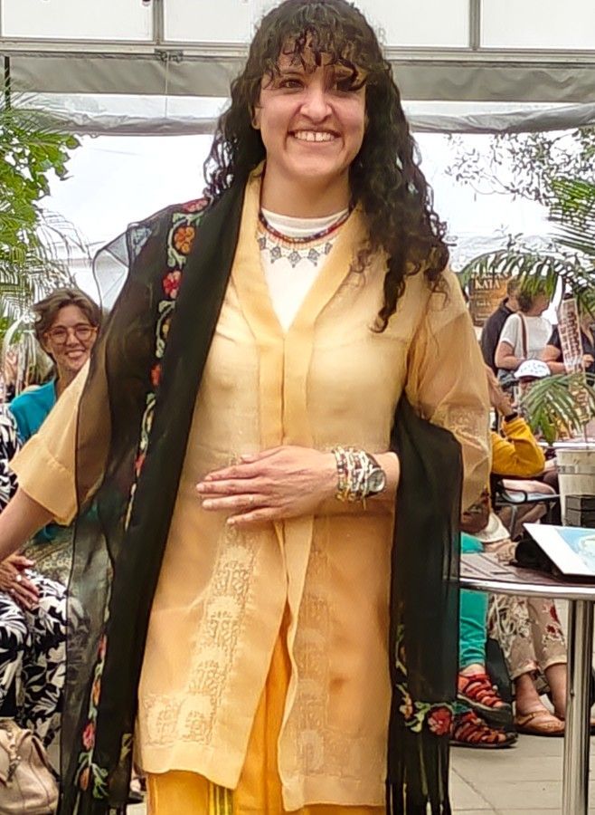 Een lachende persoon in een gouden outfit staat bij een evenement in de openlucht, met een zwarte sjaal over één schouder.