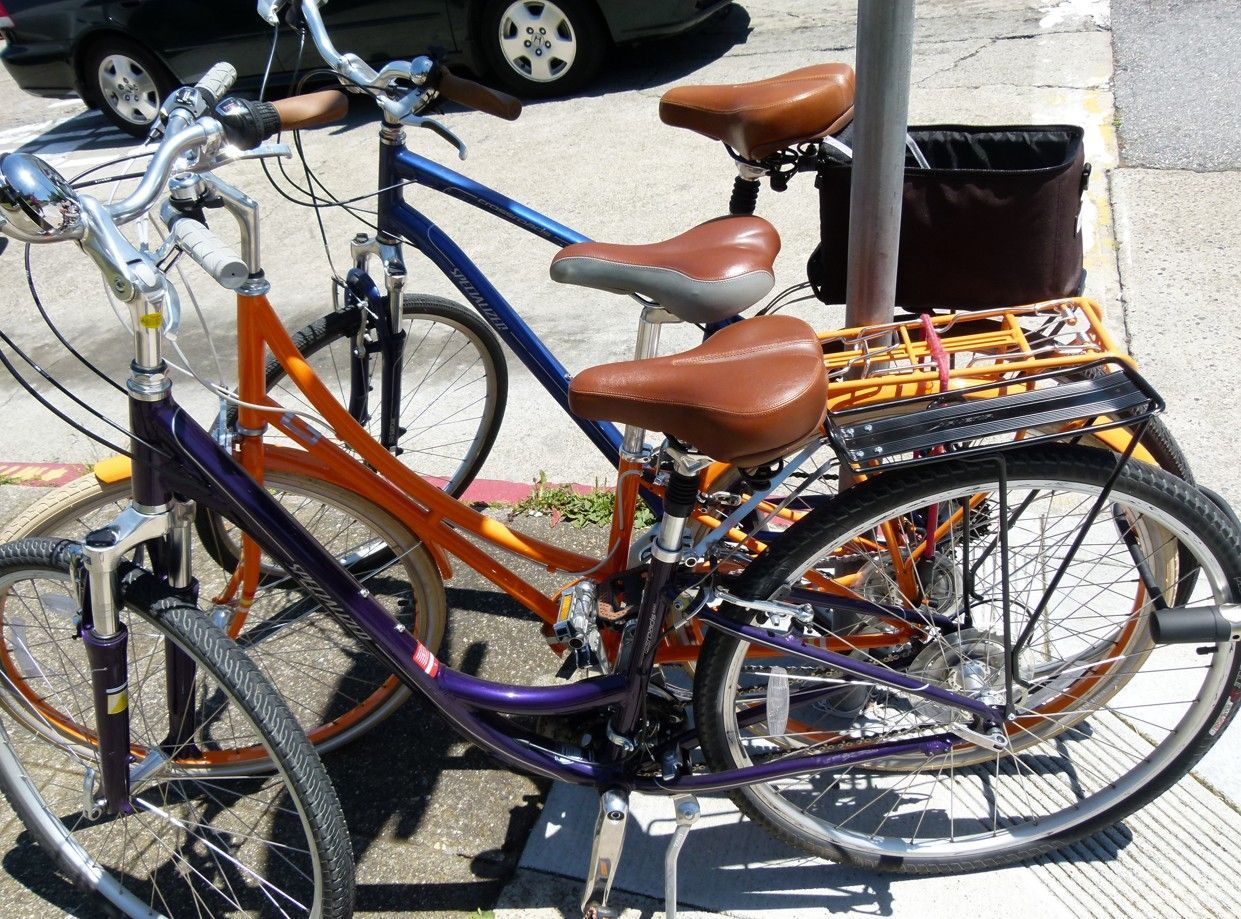 Drie fietsen aan elkaar vastgemaakt op de stoep, een paarse, een oranje en een blauwe met bruine zadels.