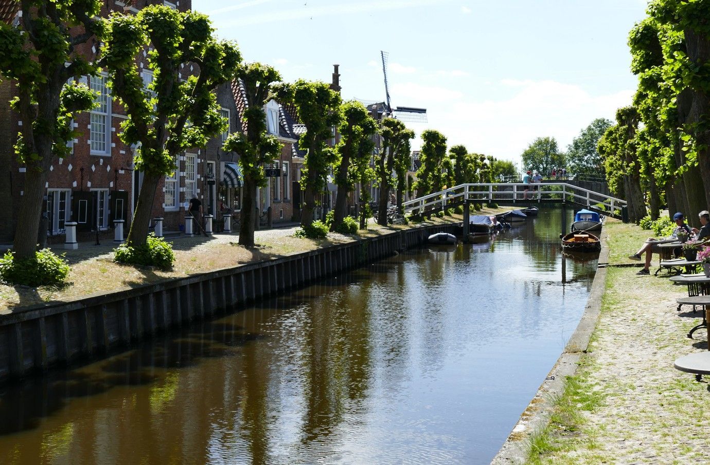 Een kanaal omzoomd met bomen en gebouwen, een kleine brug en in de verte een windmolen op een zonnige dag.