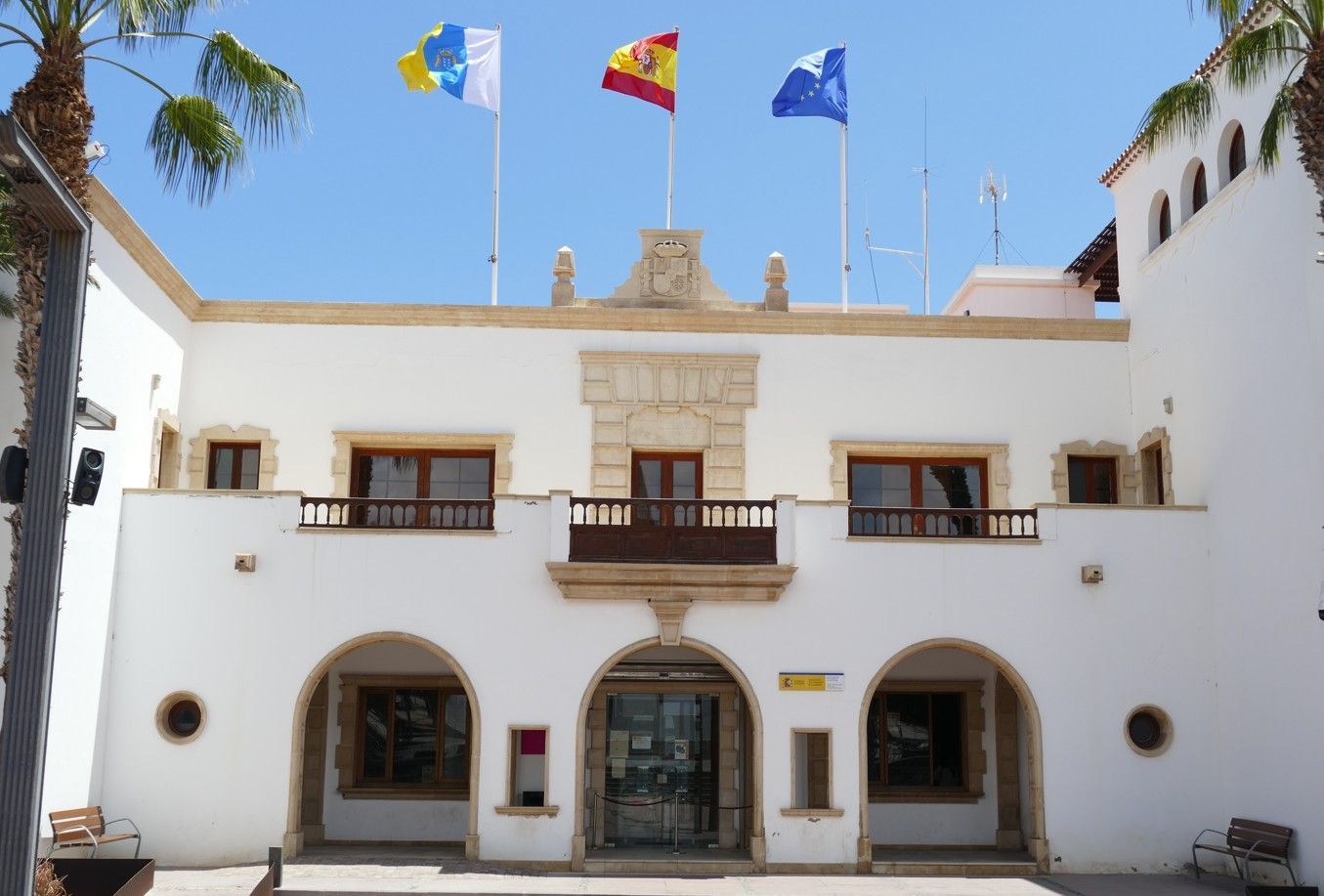 Wit gebouw met een boogvormige ingang en vlaggen van Spanje, de Canarische Eilanden en de blauwe vlag.