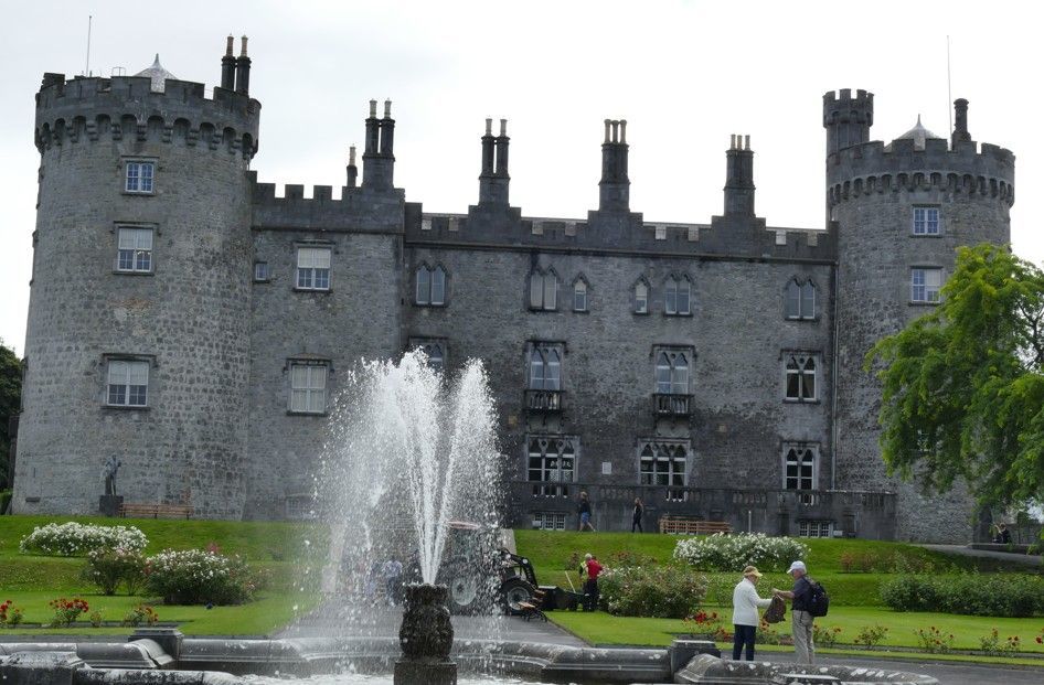 Kilkenny Castle met fontein op de voorgrond. Grijs stenen gebouw, twee ronde torens, mensen.
