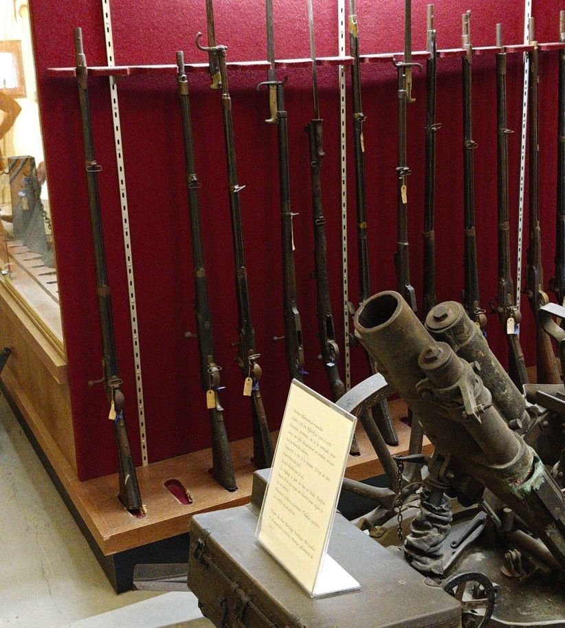 Museumtentoonstelling van oude geweren op rode wandrekken naast een opgesteld artilleriestuk.