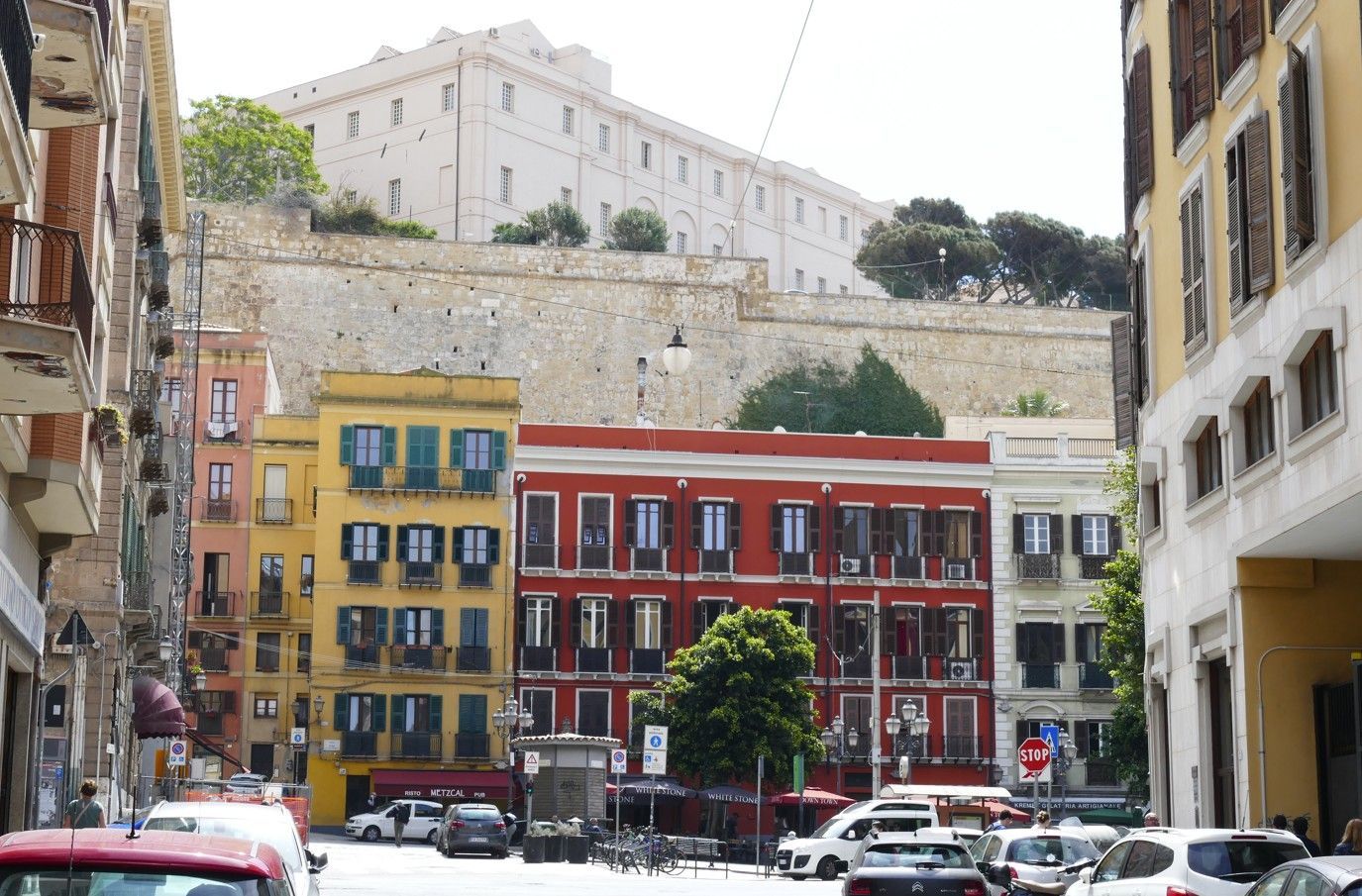 In een Italiaanse stad staan ​​kleurrijke gebouwen langs een straat, met op de achtergrond een groot fort op een heuvel.