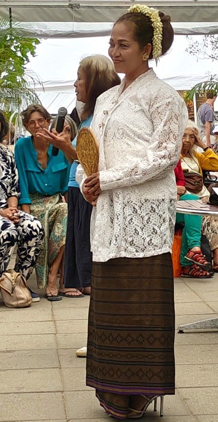Vrouw in een witte kanten blouse en een donkere batikrok staat bij een evenement in de openlucht en houdt een waaier vast.