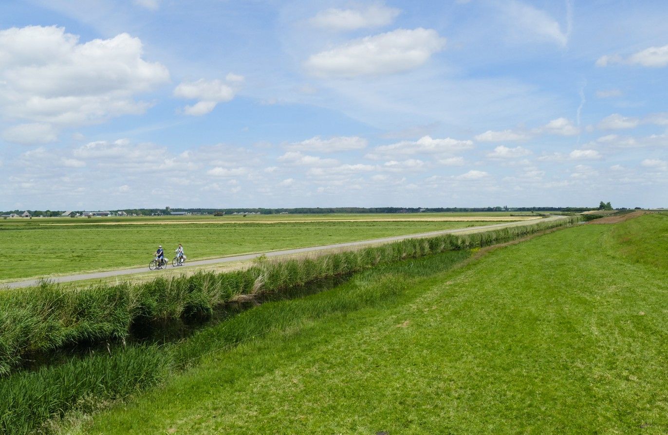 Twee fietsers op een graspad door een groen veld onder een blauwe hemel met wolken.