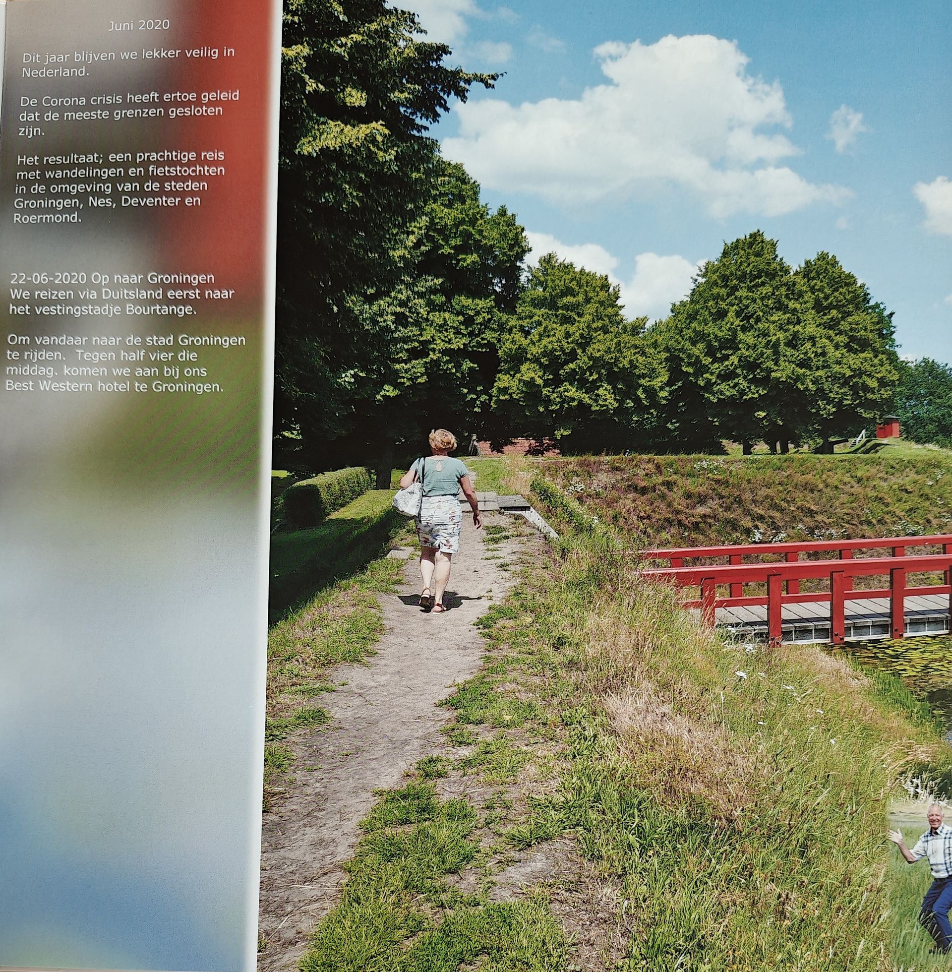 Een persoon loopt over een zandpad richting een rode brug en groene bomen. Blauwe lucht met wolken.