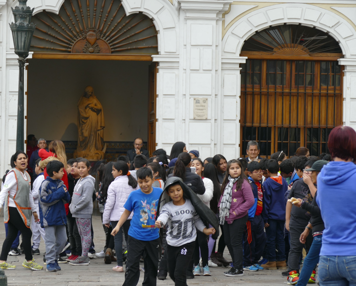 Schoolkinderen buiten een gebouw met een standbeeld, sommigen rennen, in een drukke buitenomgeving.