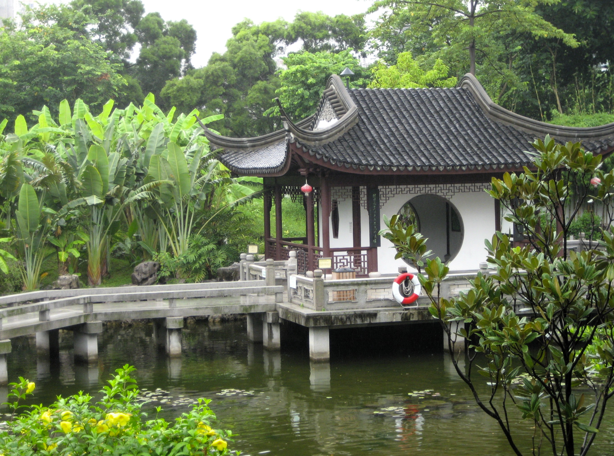Traditioneel Chinees paviljoen boven een vijver, grijs dak, witte muren, weelderig groen.