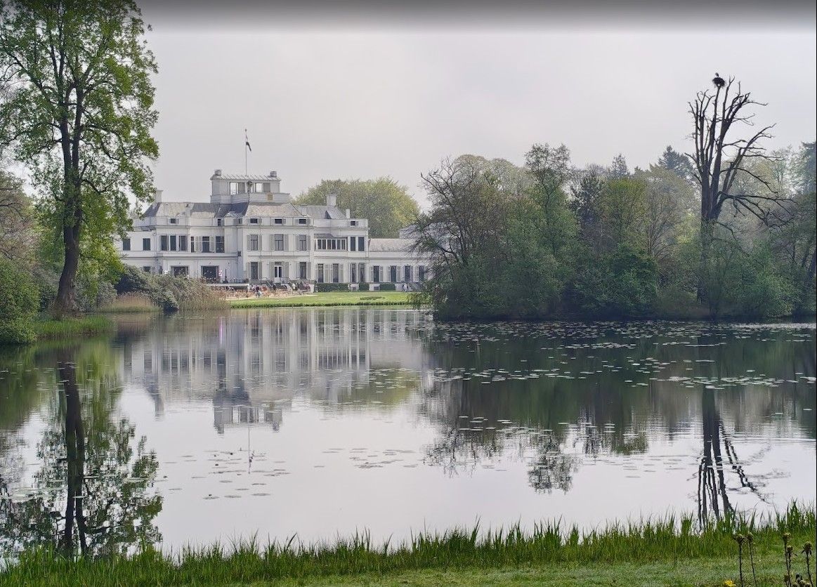 Groot, wit, statig huis aan het meer, met bomen en de lucht weerspiegeld in het kalme water.