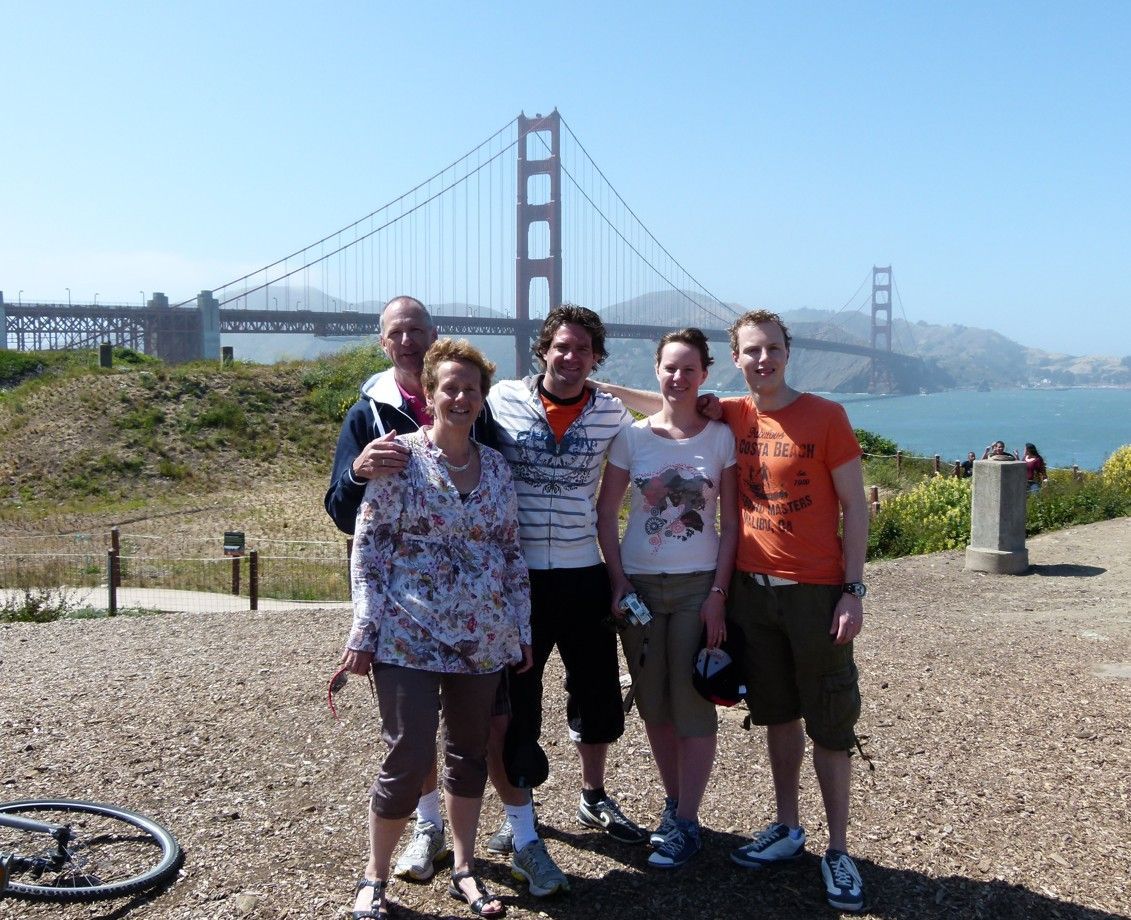 Vijf mensen poseren op een zonnige dag bij de Golden Gate Bridge. Ze glimlachen, met de brug op de achtergrond.