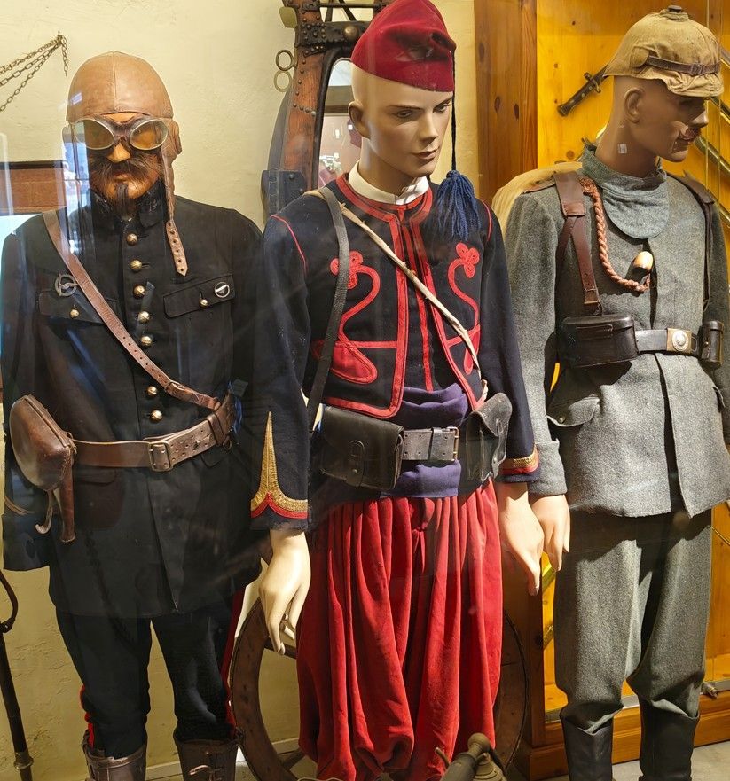 Drie mannequins in traditionele kostuums, in zwarte, rode en grijze outfits, staan ​​binnen tentoongesteld.