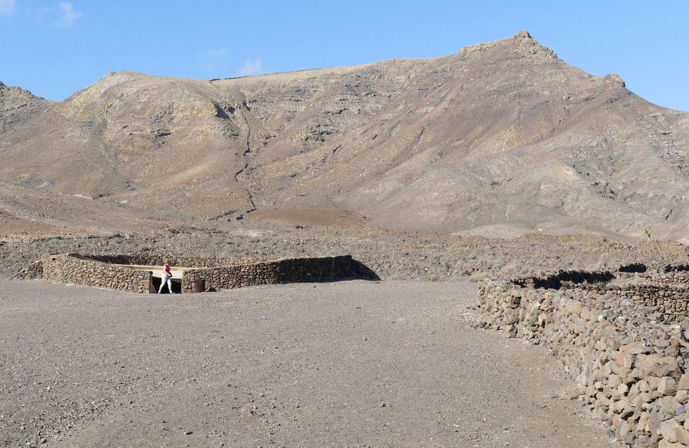 Een stenen constructie en muren in een kaal landschap, met een berg op de achtergrond. 