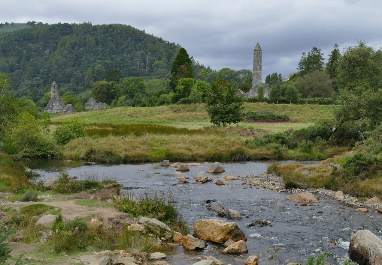 De rivier stroomt door een groen landschap richting een oude stenen toren en ruïnes onder een bewolkte hemel.