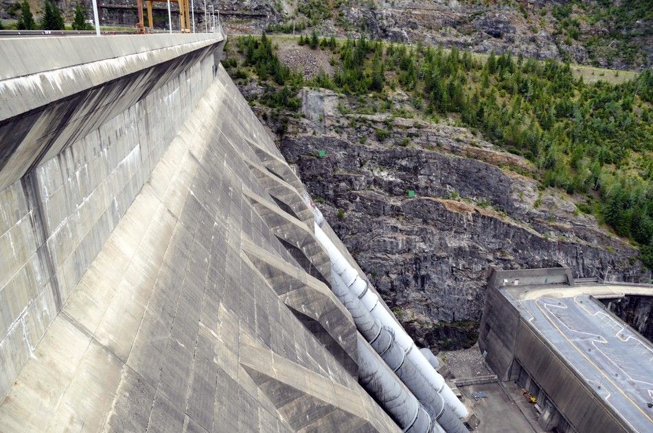 Een betonnen dam met diagonale buizen tegen een rotsachtige helling en enkele bomen.