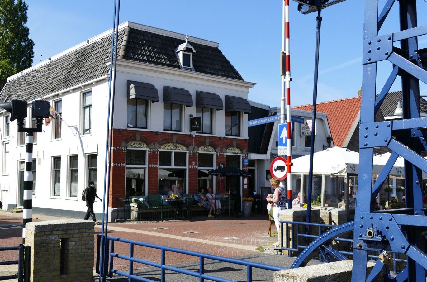 Een wit gebouw van twee verdiepingen met een luifel, terras en een blauwe brug. Een spoorwegovergang blokkeert de toegang.